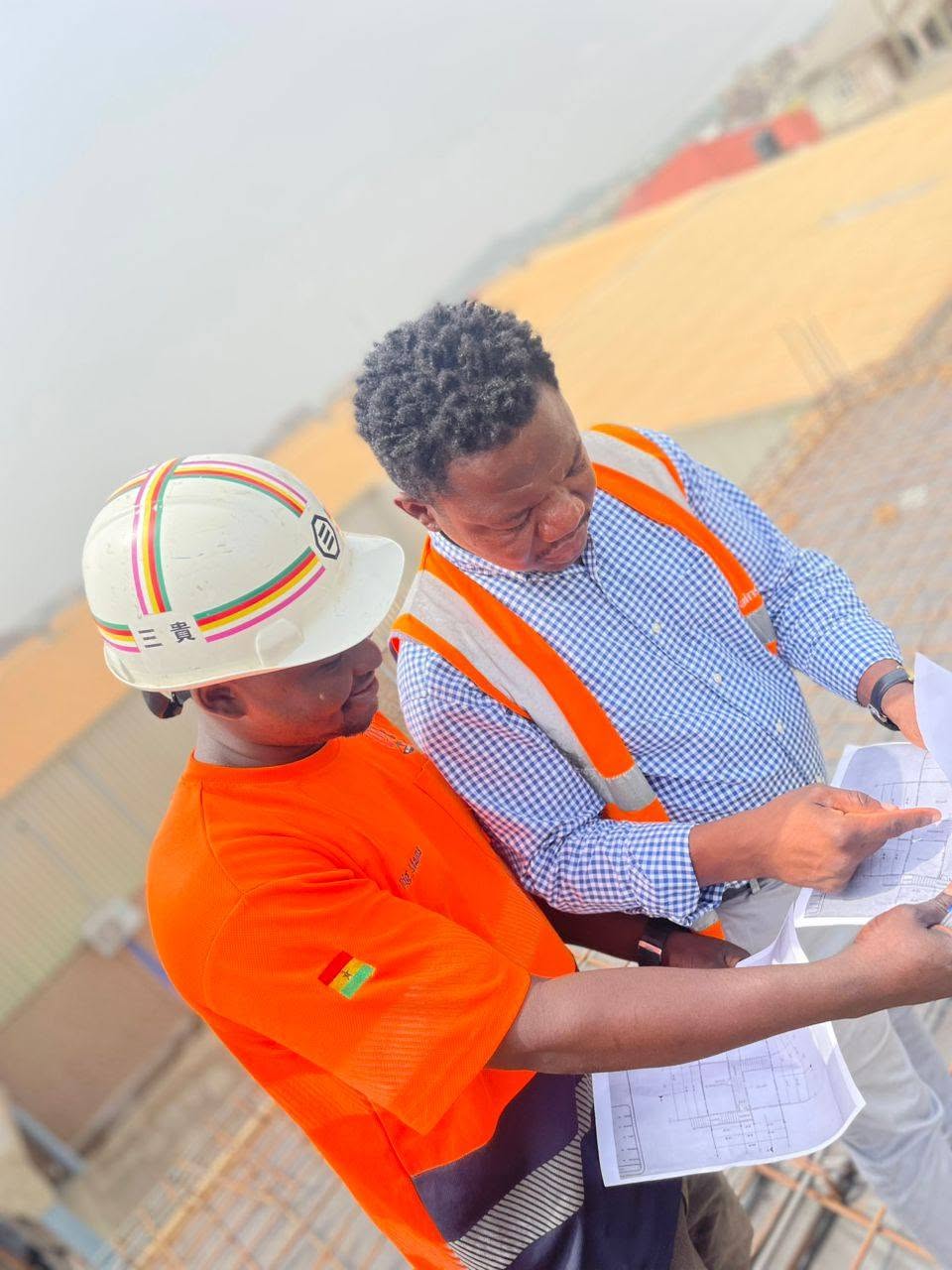 X-Mell engineer in safety helmet and high-visibility vest reviewing construction drawings with a colleague on site